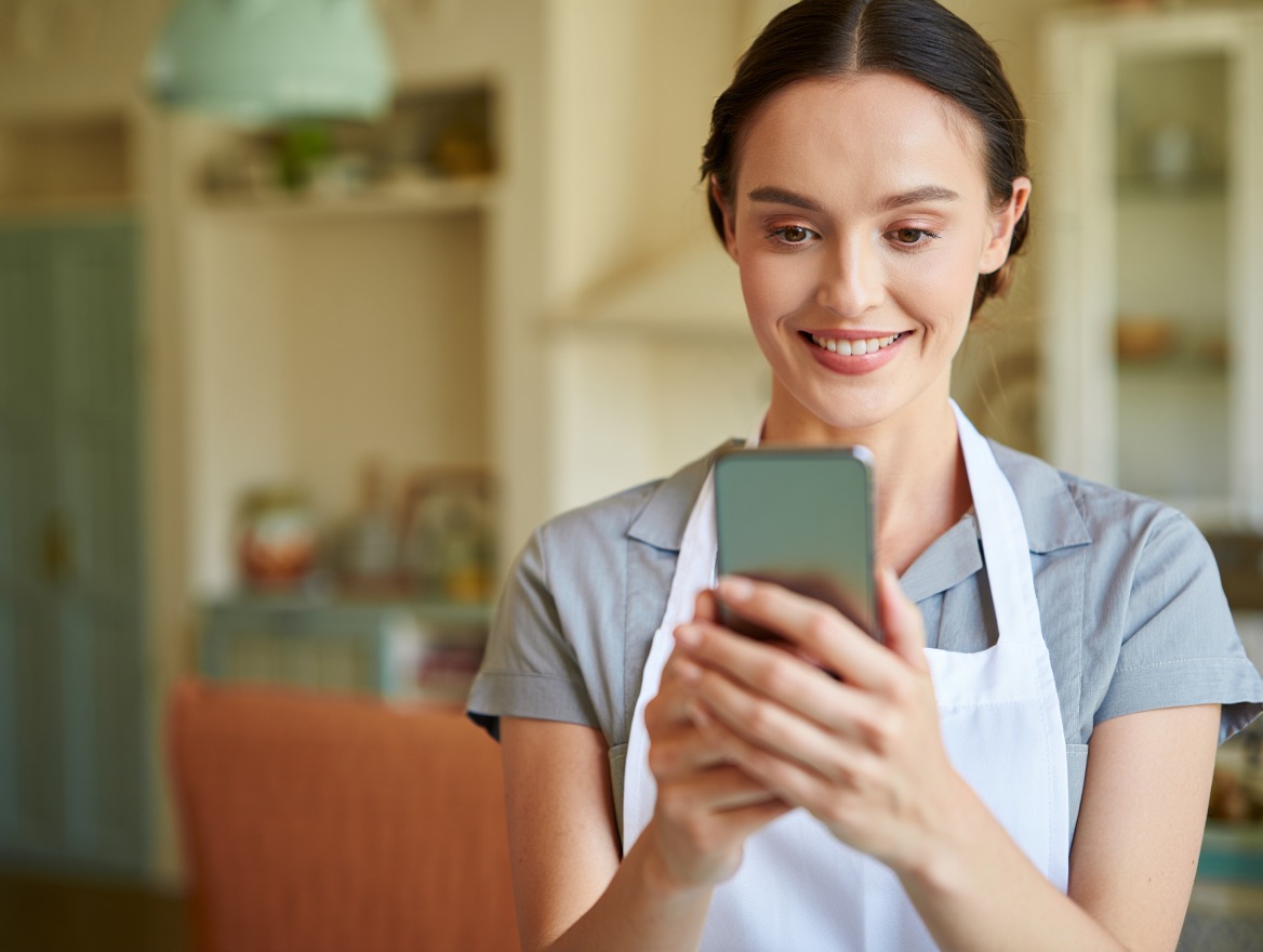 Empregada doméstica sorrindo na cozinha enquanto bate o ponto pelo celular